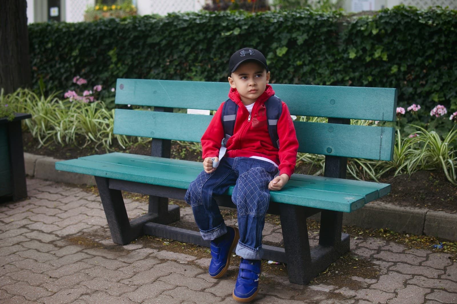 Un garçon en sweat à capuche rouge et casquette, assis sur un banc vert.