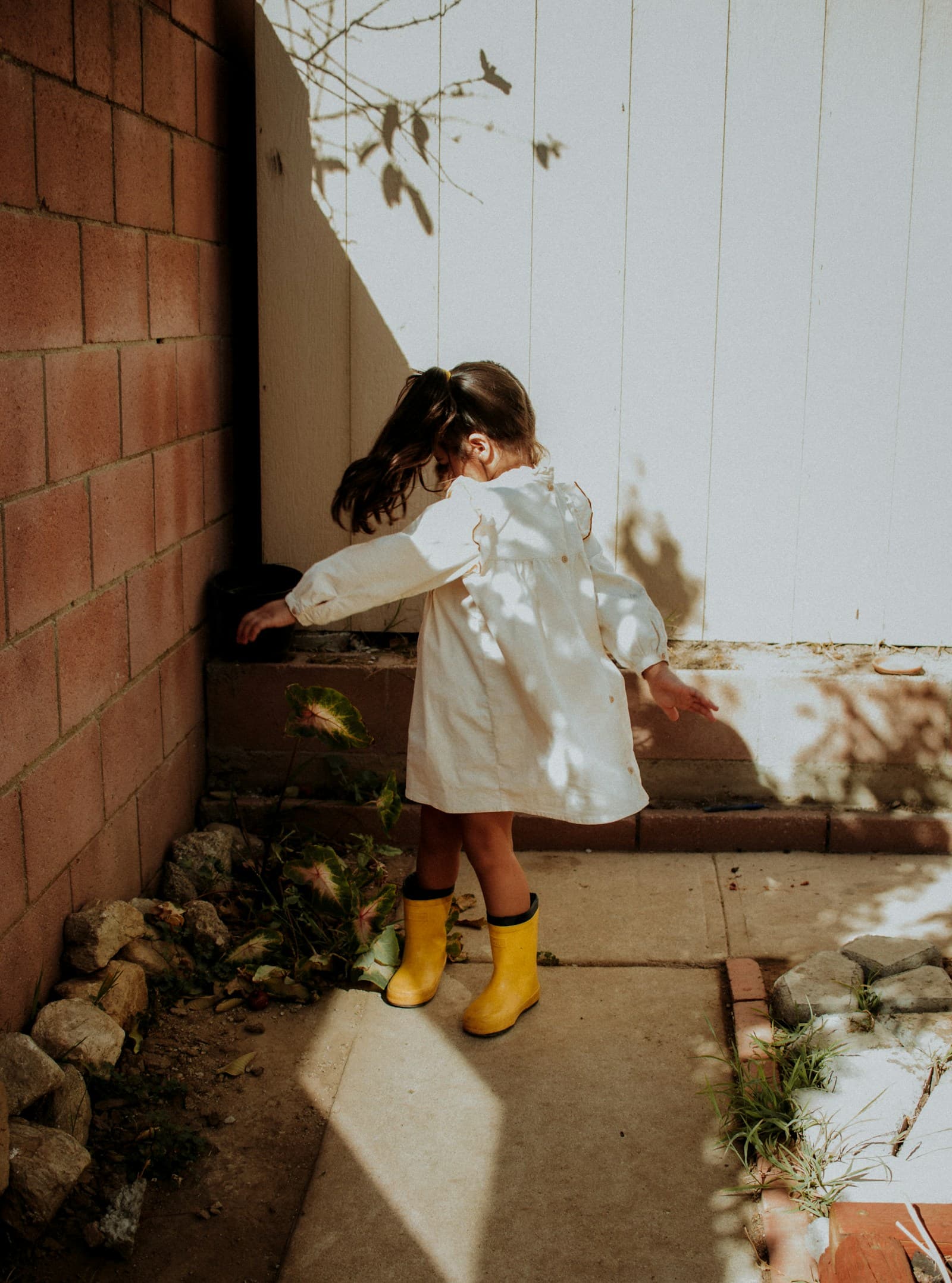 Une fille en manteau blanc et bottes jaunes, devant un mur de briques.